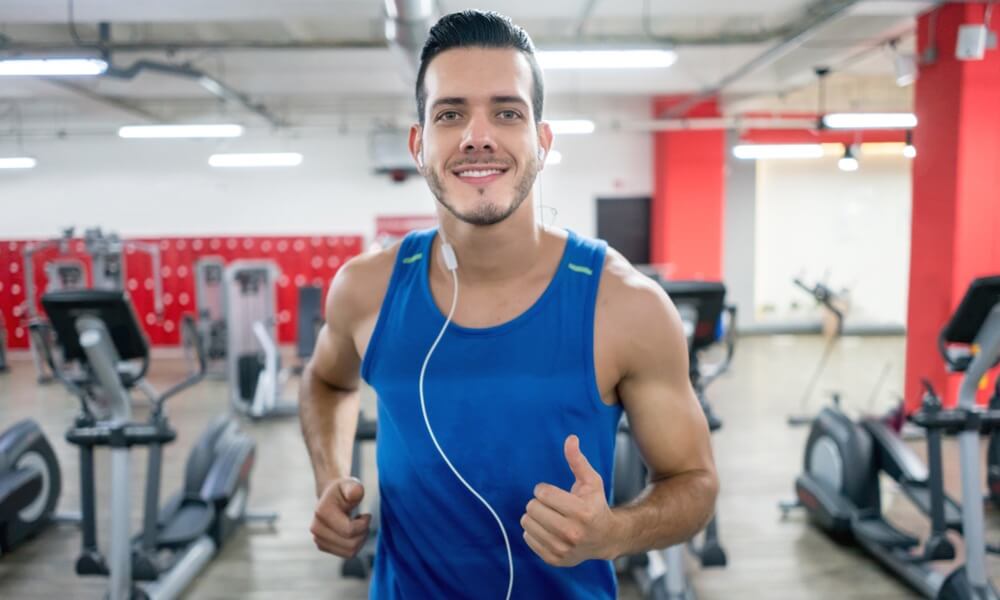 Ein Mann im blauen Shirt läuft auf einem Laufband im Fitnessstudio und lächelt in die Kamera