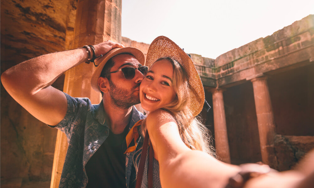 Ein junges Ehepaar in Freizeitkleidung macht ein Selfie vor einer historischen Tempelanlage.