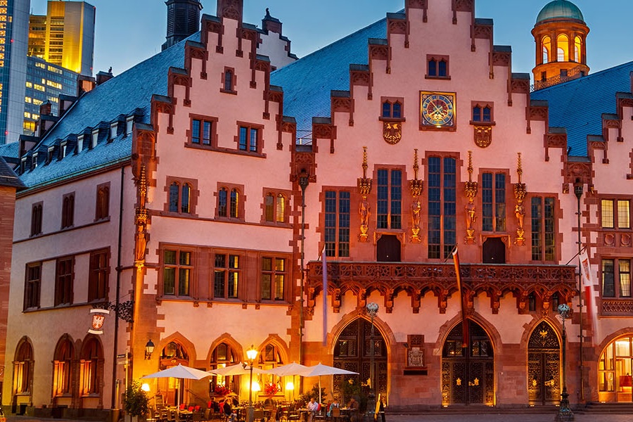 Frankfurt_Städteseiten Das Foto zeigt den Römerplatz in Frankfurt am Main bei Dämmerung. Im Vordergrund sind historische Gebäude mit beleuchteten Fenstern zu sehen. Im Hintergrund ragt ein modernes Hochhaus in den Himmel.