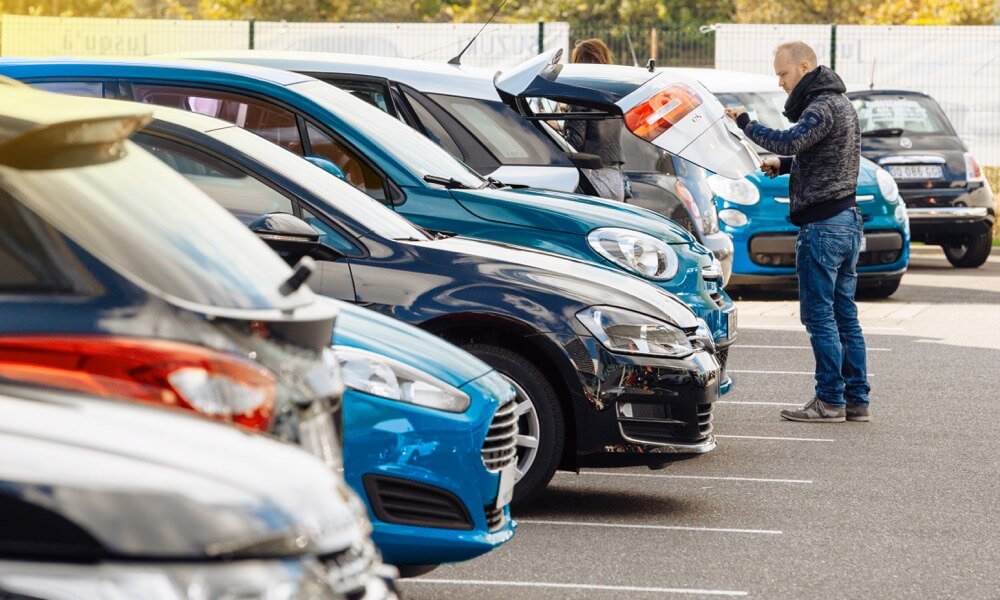 Ein Mann öffnet des Kofferraum eines Autos. Daneben stehen weitere Autos.