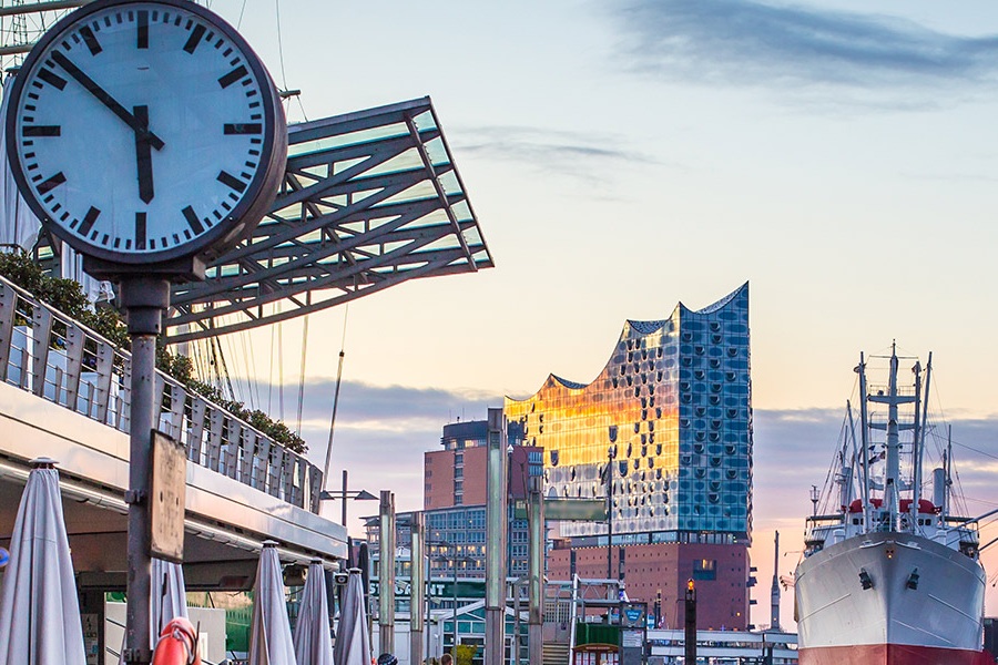 Foto von Hamburg an der Elbe bei Dämmerung. Im Vordergrund ist eine Promenade mit geschlossenen Sonnenschirmen und einer großen analogen Uhr. Im Hintergrund sind ein großes Schiff und die Elbphilharmonie zu sehen.