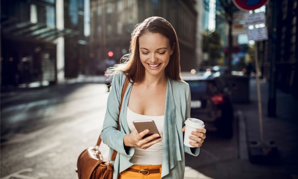 Frau geht lächelnd den Gehweg entlang, schaut auf ihr Smartphone und hält in der anderen Hand einen ToGo-Becher.