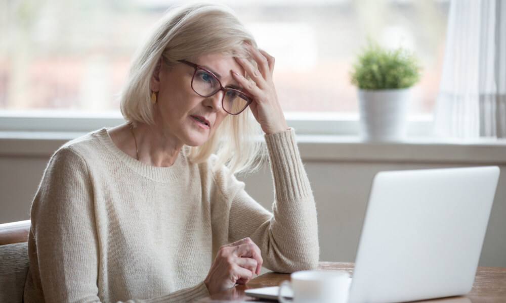 Eine ältere Frau sitzt vor einem Laptop, sie fasst sich an die Stirn und hat einen besorgten Gesichtsausdruck.