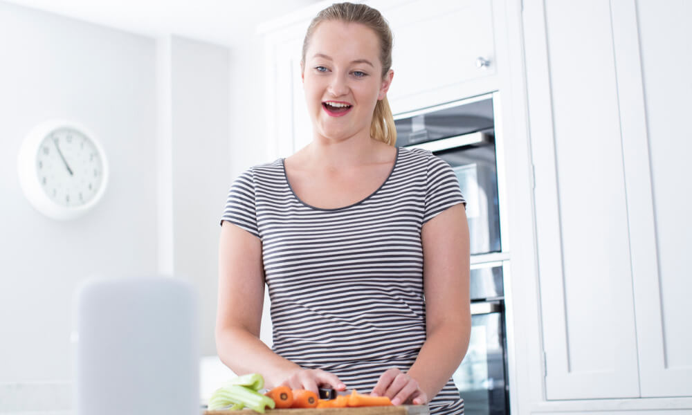 Eine Frau schneidet in einer Küche Gemüse und spricht dabei mit einem Smart Speaker.