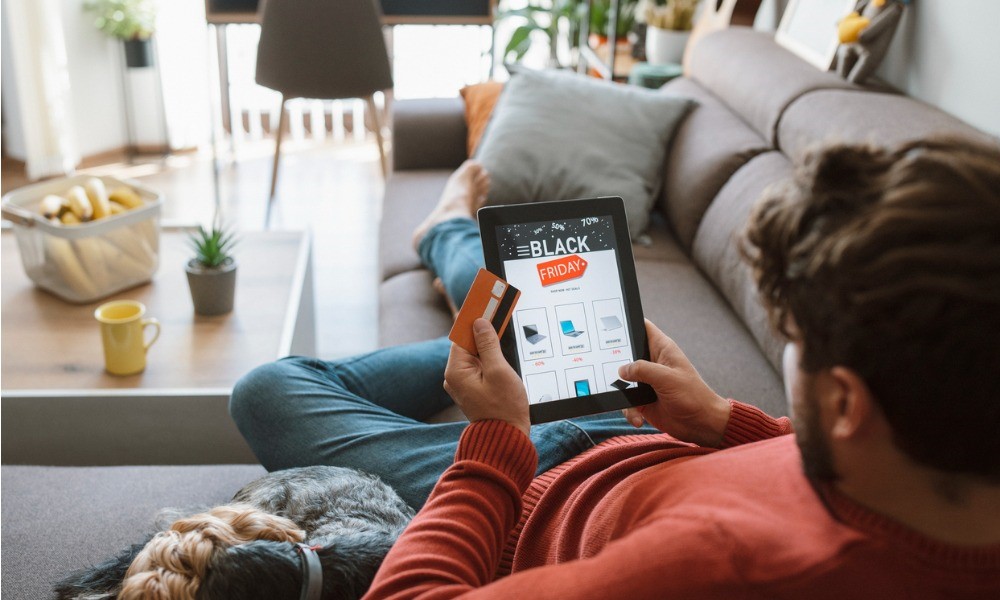 Mann liegt mit Hund zuhause auf dem Sofa und shoppt am Black Friday online am Tablet.