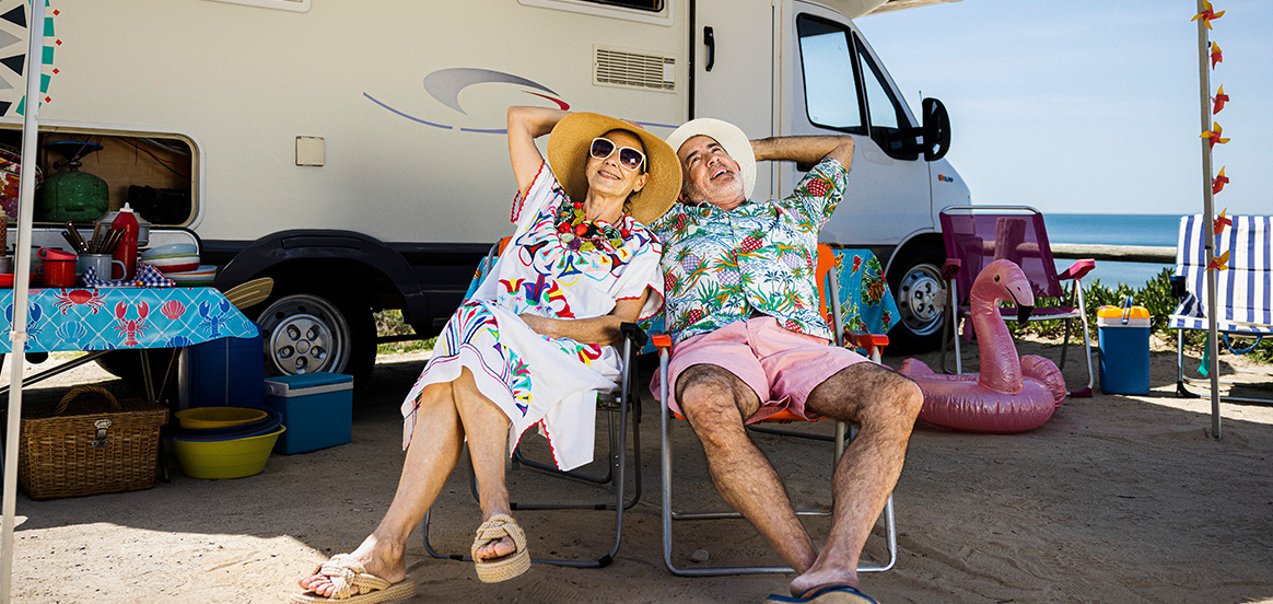Eine Frau und ein Mann sitzen in Campingstühlen vor ihrem Wohnmobil. Sie tragen Sonnenhüte und bunte Sommerkleidung. Das Wohnmobil steht auf Sand mit Blick aufs Meer. Um das Wohnmobil liegen Camping- und Strandutensilien wie ein Eiskühler, Geschirr und ein pinker Flamingo-Schwimmring.