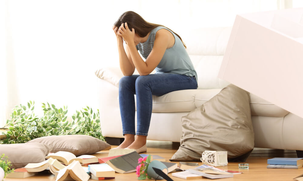 Eine Frau sitzt auf dem Sofa und rauft sich die Haare. Das Zimmer, in dem sie sich befindet, ist verwüstet. Gegenstände liegen wild auf dem Fußboden verstreut.