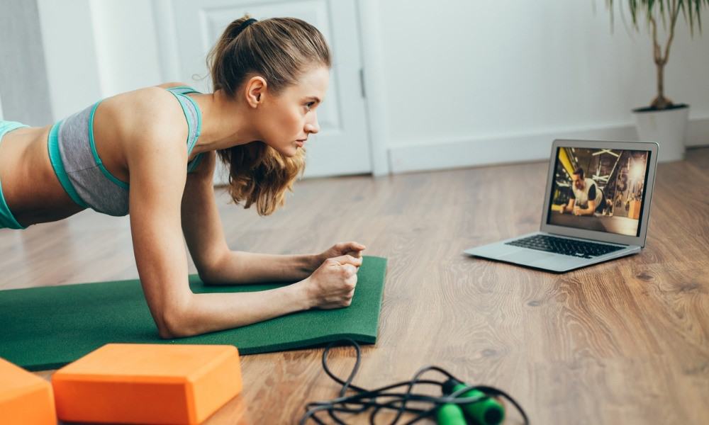 Frau übt eine Plank auf einer Matte vor ihrem Laptop auf dem Boden im Wohnzimmer aus.