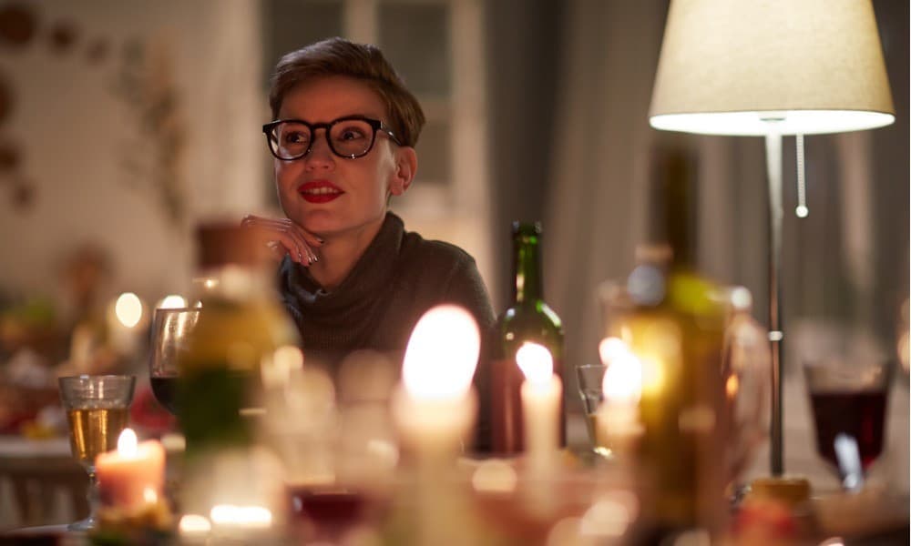 Frau mit Brille sitzt in gemütlicher Atmosphäre bei Kerzenschein und Tischleuchte am gedeckten Tisch.