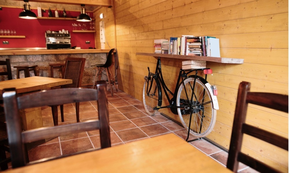 Ein Damenrad lehnt in einem Restaurant an einer Holzwand. Parallel zur Wand liegt ein Holzbrett auf dem Rad. Auf diesem stehen Bücher.