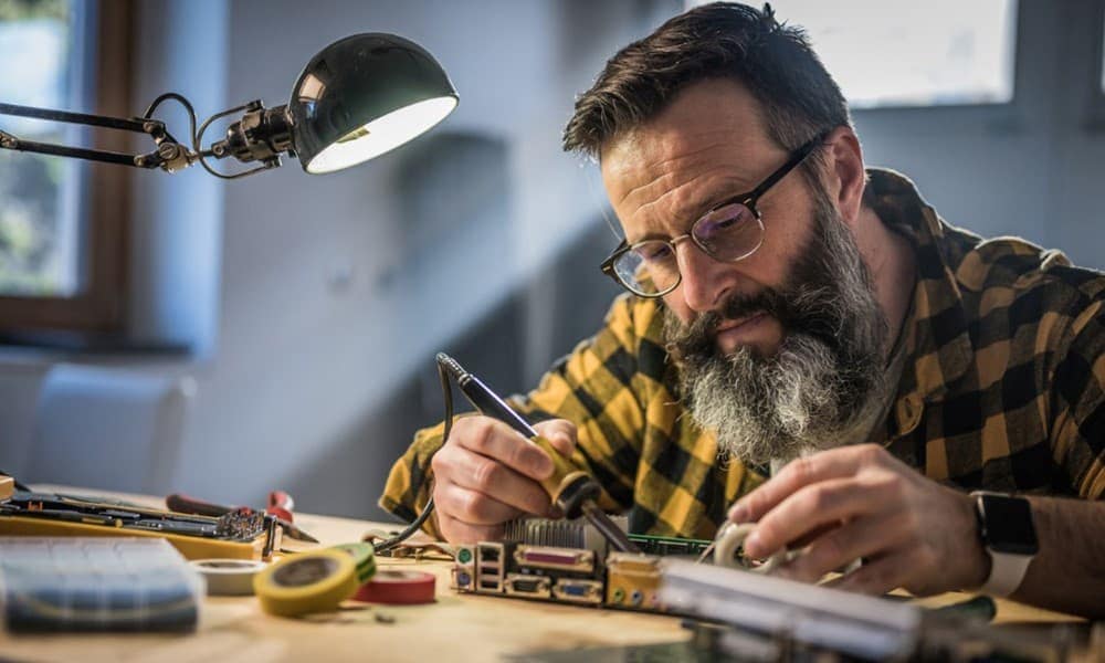 Nahaufnahme eines Technikers, der einen Lötkolben benutzt, um eine Platine in der Werkstatt unter einer Schreibtischlampe zu reparieren.