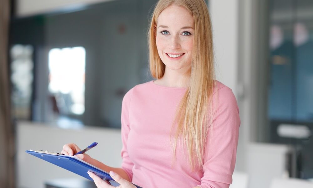 Eine junge fRau im rosa Pullover lächelt in die Kamera und hält Stift und Klemmboard in der Hand.